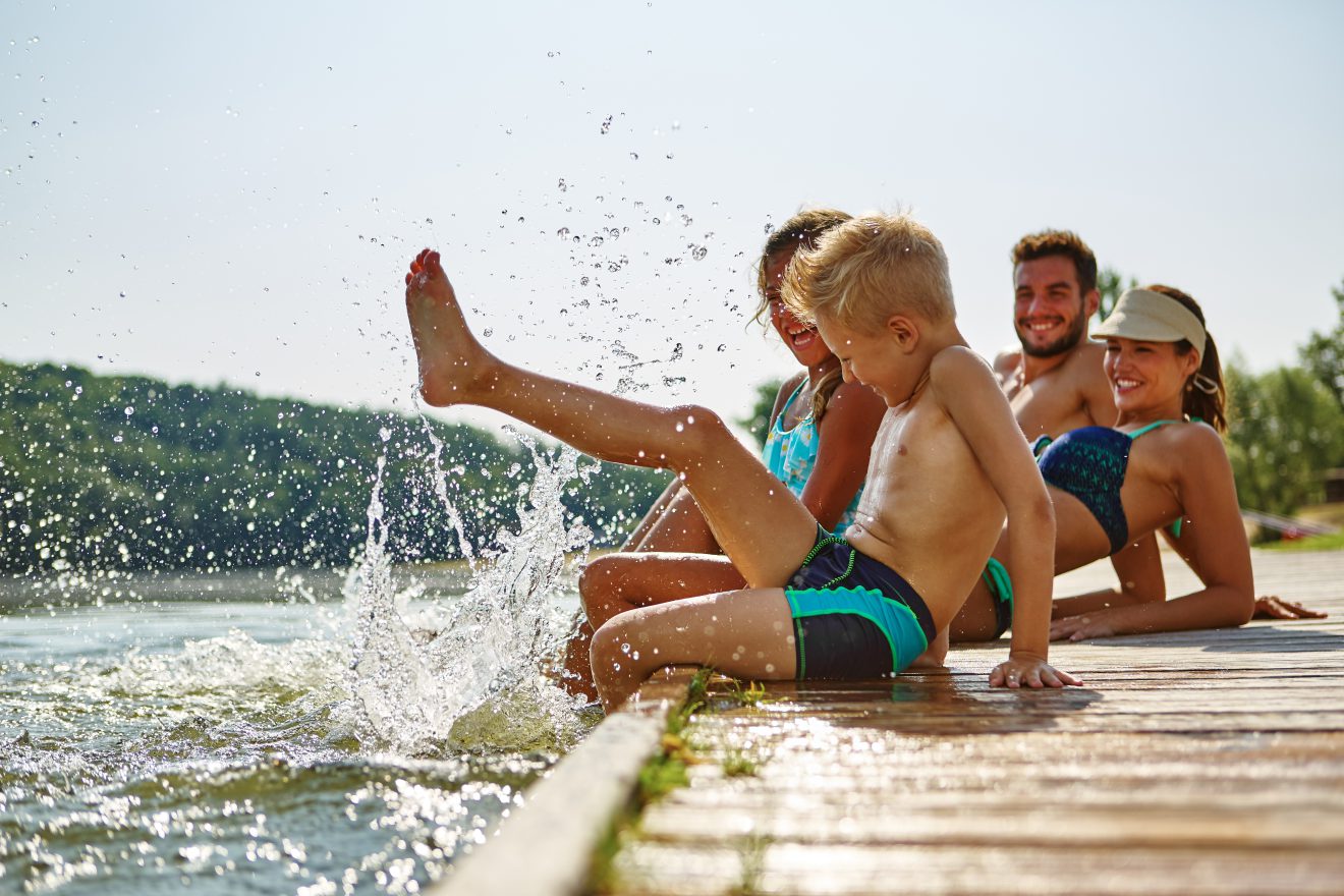 Familie am See badet glücklich mit Füßen im Wasser im Sommer. Symbolbild für sechste Urlaubswoche