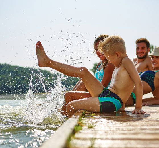 Familie am See badet glücklich mit Füßen im Wasser im Sommer. Symbolbild für sechste Urlaubswoche