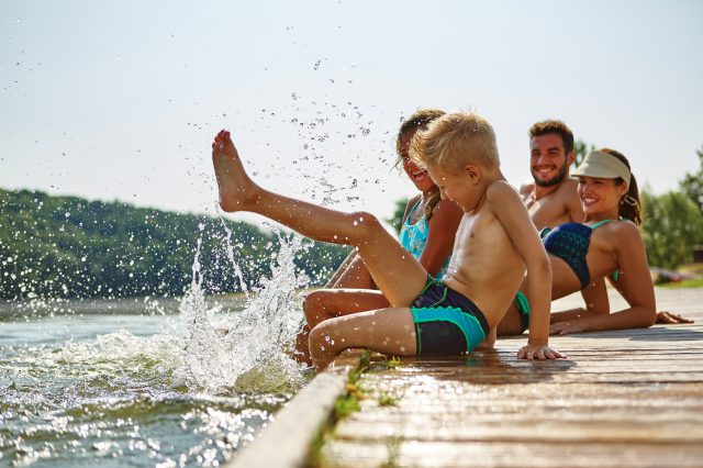 Familie am See badet glücklich mit Füßen im Wasser im Sommer. Symbolbild für sechste Urlaubswoche