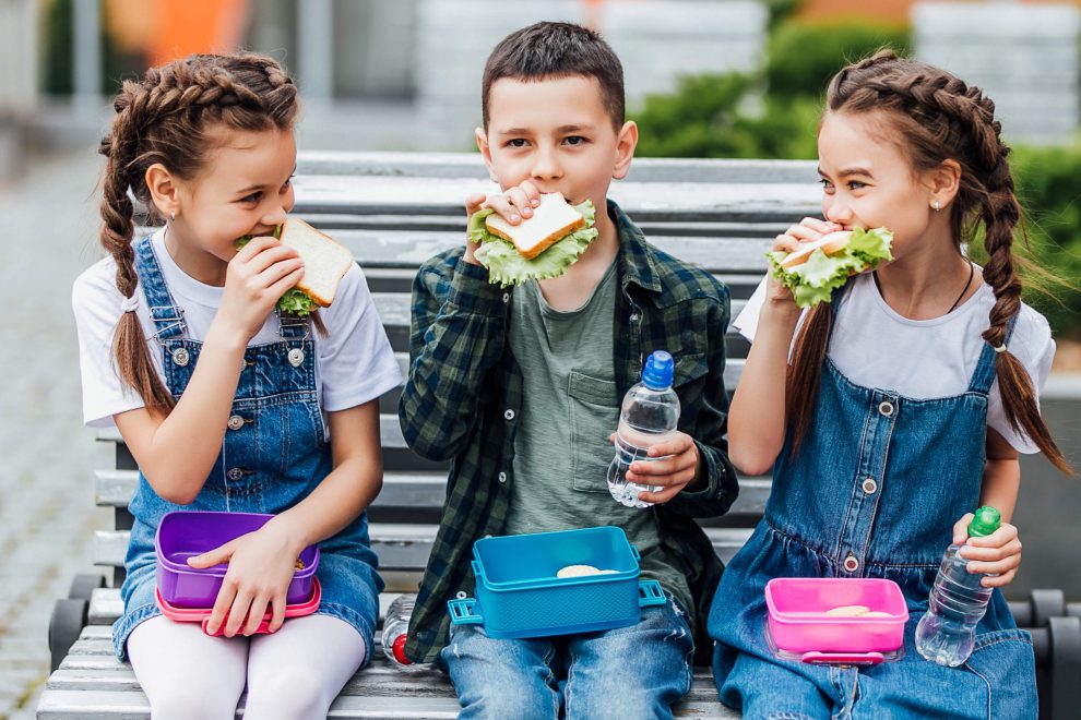Kinder sitzen auf einer Bank und essen ihr Pausenbrot in der Schule. Die Inflation wirkt sich auch auf Kinder aus.