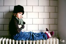 Ein Kind sitzt mit Mütze und Schal auf einem Heizkörper. Symbolbild für die Armut infolge der Teuerung, die Familien in die Armut treibt.