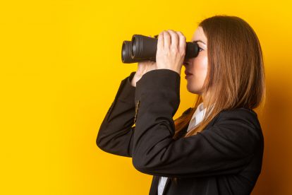 Eine Frau steht vor einer gelben Wand und blickt mit dem Fernglas zurück. Symbolbild für den Jahresrückblick 2022.