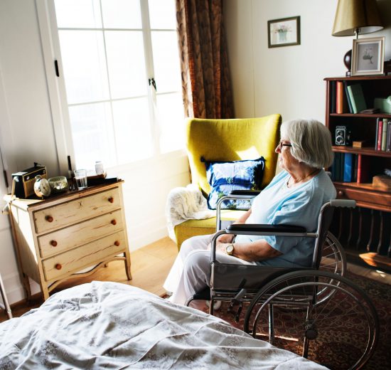 Eine ältere Frau im Rollstuhl sitzt alleine in ihrem Zimmer und wartet auf die 24-Stunden-Betreuung.