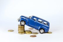 Ein Spielzeugauto ist an einem Turm auf Münzen angelehnt. Symbolbild für die unleistbaren Alltagskosten.