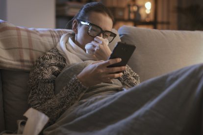 Eine Frau hat eine Erkältung und liegt mit dem Handy in der Hand auf der Couch. Symbolbild für krank in die Arbeit.