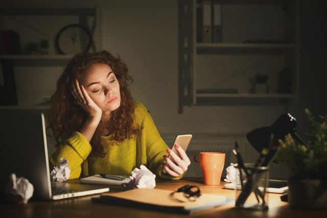 Eine Frau sitzt vor ihrem Laptop und schaut müde auf ihr Handy. Symbolbild für die ständige Erreichbarkeit, mit der Beschäftigte umgehen müssen.