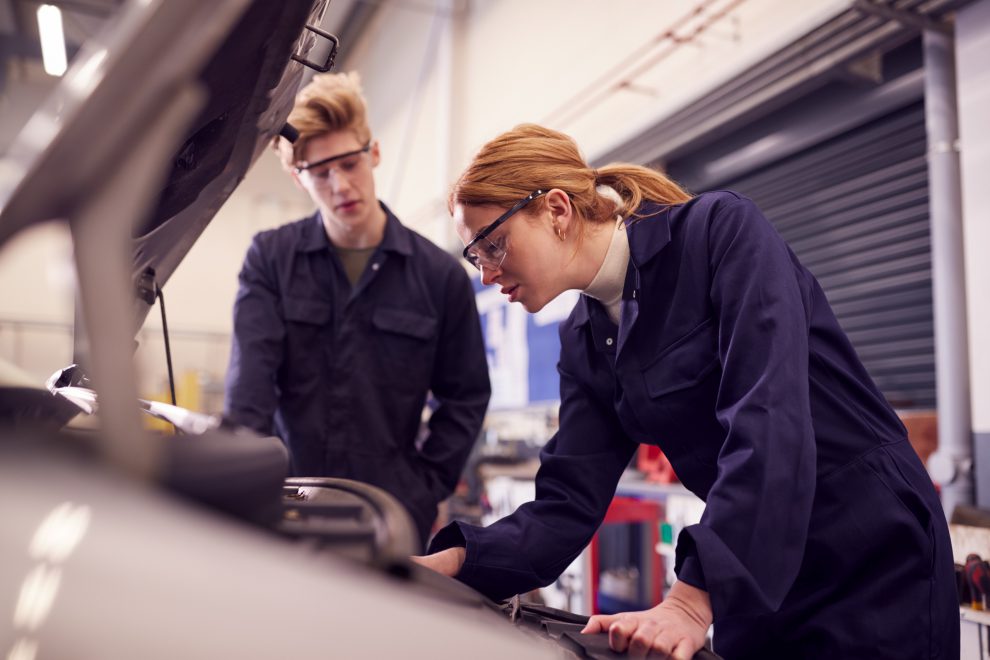 Eine junge Frau repariert ein Auto. Im Hintergrund steht ein junger Mann. Symbolbild für den Arbeitsdruck, den junge Menschen erleben.