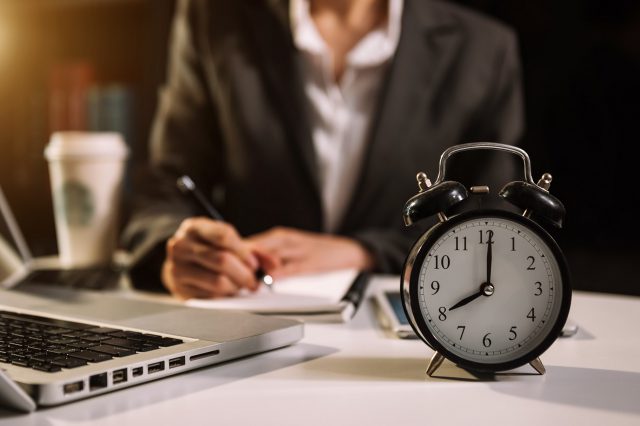 Eine Frau sitzt an ihrem Schreibtisch, vor ihr ein Laptop und ein Wecker. Symbolbild für die Arbeitszeitverkürzung.