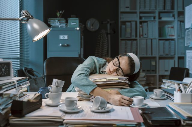 Eine Frau schläft in einem Büro auf Aktenordnern, vor sich mehrere Tassen Kaffee. Symbolbild für den hohen Arbeitsdruck und die sinkende Arbeitszufriedenheit.