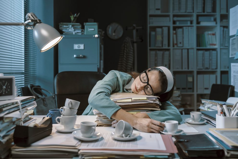 Eine Frau schläft in einem Büro auf Aktenordnern, vor sich mehrere Tassen Kaffee. Symbolbild für den hohen Arbeitsdruck und die sinkende Arbeitszufriedenheit.