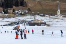 Ein Skiort mit nur leicht beschneiter Piste. Im Hintergrund ist ein Skilift zu sehen, im Vordergrund drei Menschen auf Skiern. Symbolbild für den Wintertourismus und seine Gefährdung.