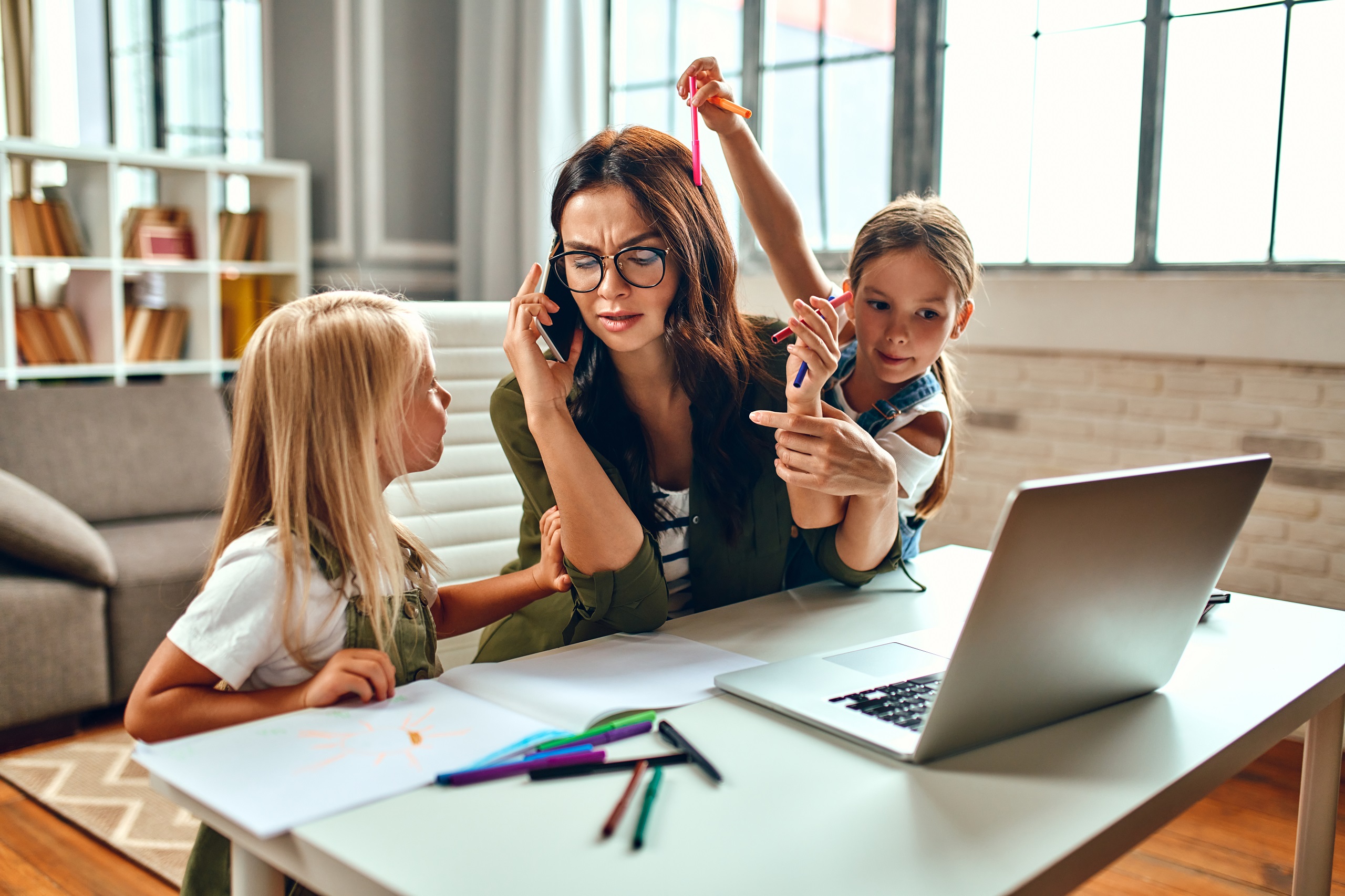 Eine Frau spricht in ein Telefon und arbeitet an einem Laptop. Zwei Kinder buhlen um ihre Aufmerksamkeit. Symbolbild für den Spagat zwischen Familie und Beruf.