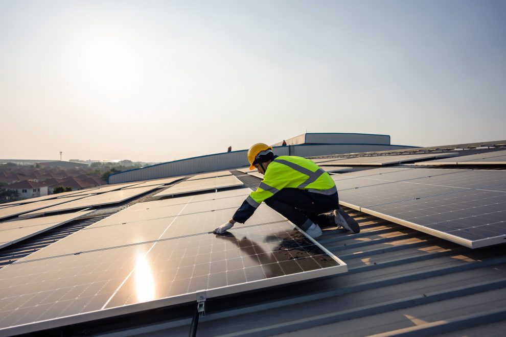 Ein Mann in Schutzkleidung steht auf einem Hausdach und arbeitet an einer Fotovoltaikanlage. Symbolbild für Green Jobs.