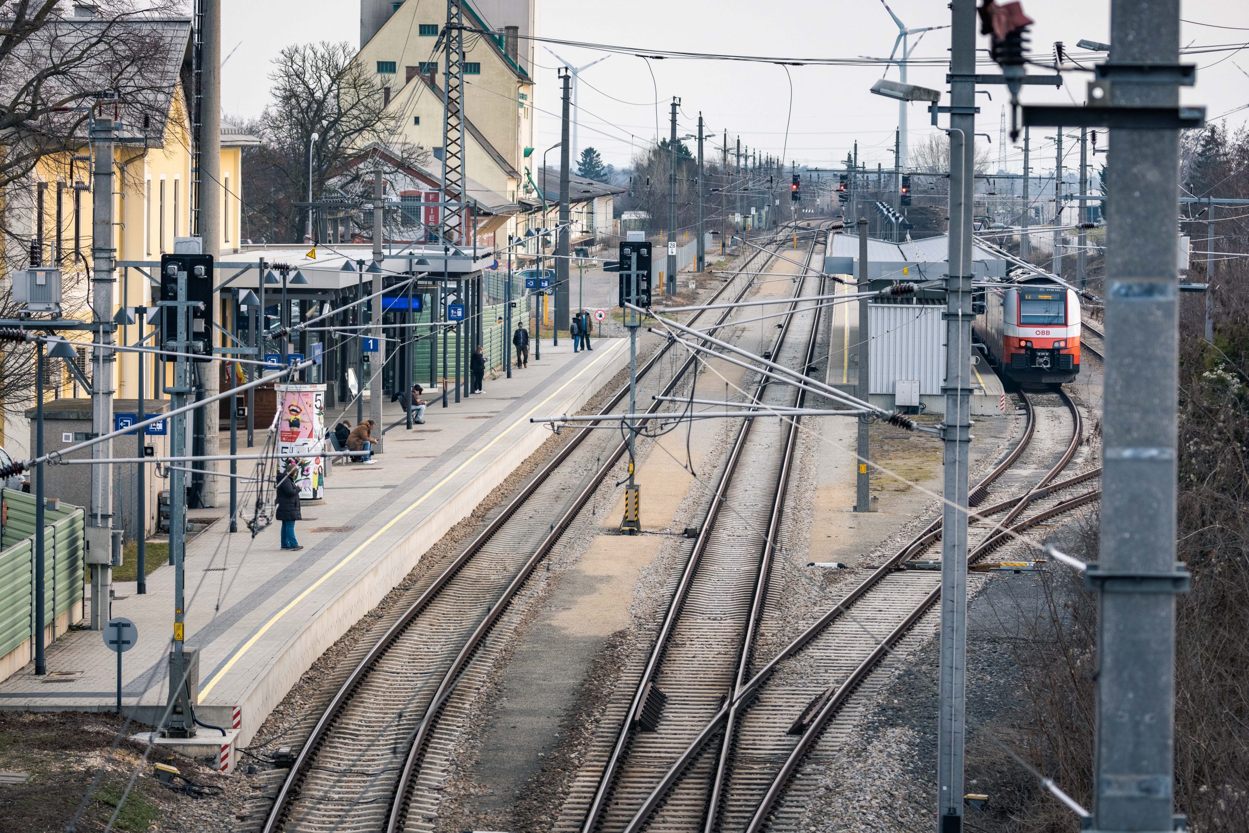 Ein Bahnhof, fotografiert von einer Brücke. Auf dem Gleis stehen Menschen, eine Anzeige ist zu sehen und ein Zug fährt ein. Symbolbild für klimafittes Pendeln.