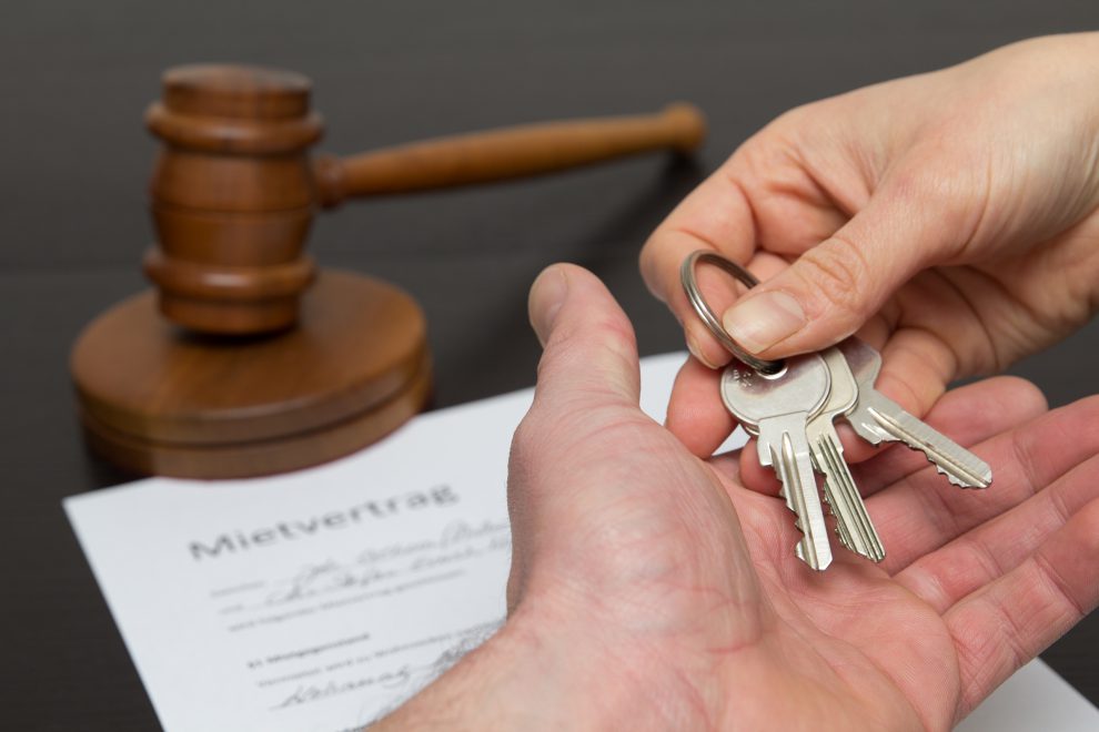 Eine Schlüsselübergabe für eine Mietwohnung. Im Hintergrund ist ein Richterhammer und ein Mietvertrag zu sehen, vorne reicht eine Hand einer anderen einen Schlüssel. Symbolbild für das Mietrecht.