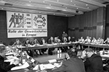 Männer sitzen an quadratisch angeordneten Tischen und halten eine Sitzung ab. Im Hintergrund ein Plakat mit der Inschrift "Rolle der Gewerkschaften in Europa", der EGB lieferte den Rahmen für eine Konferenz.