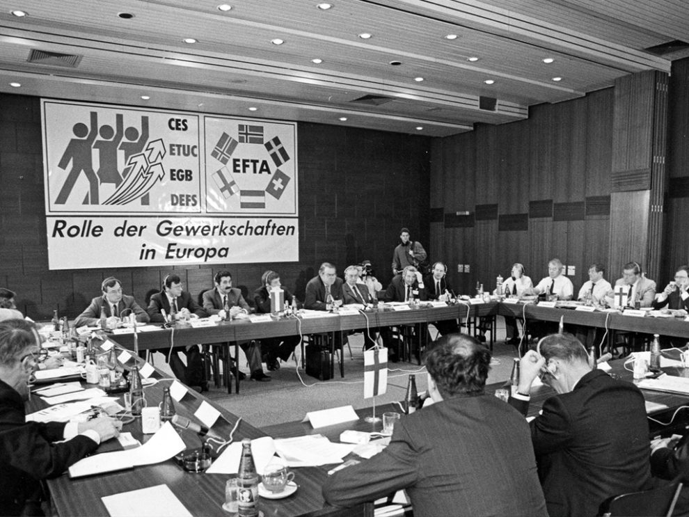 Männer sitzen an quadratisch angeordneten Tischen und halten eine Sitzung ab. Im Hintergrund ein Plakat mit der Inschrift "Rolle der Gewerkschaften in Europa", der EGB lieferte den Rahmen für eine Konferenz.