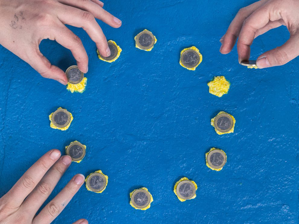 Bild von einem blauen Kuchen mit gelben Sternen, der die EU-Flagge darstellen soll. Hände legen 1-Euro-Münzen auf die Sterne. Symbolbild für die Fiskalregeln.