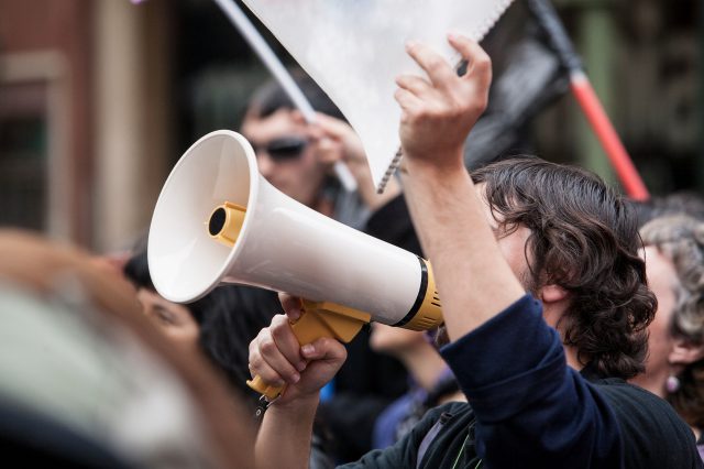 Ein Mann ist auf einer Demonstration und redet in ein Megaphon. Er hält einen Block.