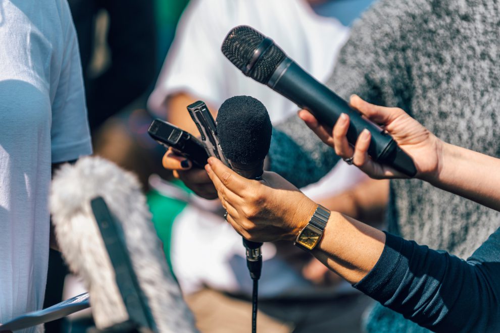 Menschen mit Mikrofonen und Diktiergeräten in der Hand interviewen eine Person. Symbolbild für die schwierigen Arbeitsbedingungen von Journalist:innen.