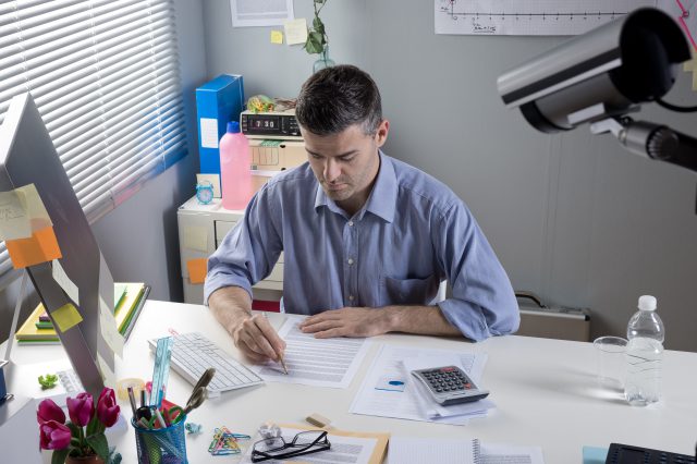 Ein Mann sitzt an einem Schreibtisch und arbeitet. Eine Überwachungskamera beobachtet ihn. Symbolbild für Überwachung am Arbeitsplatz.