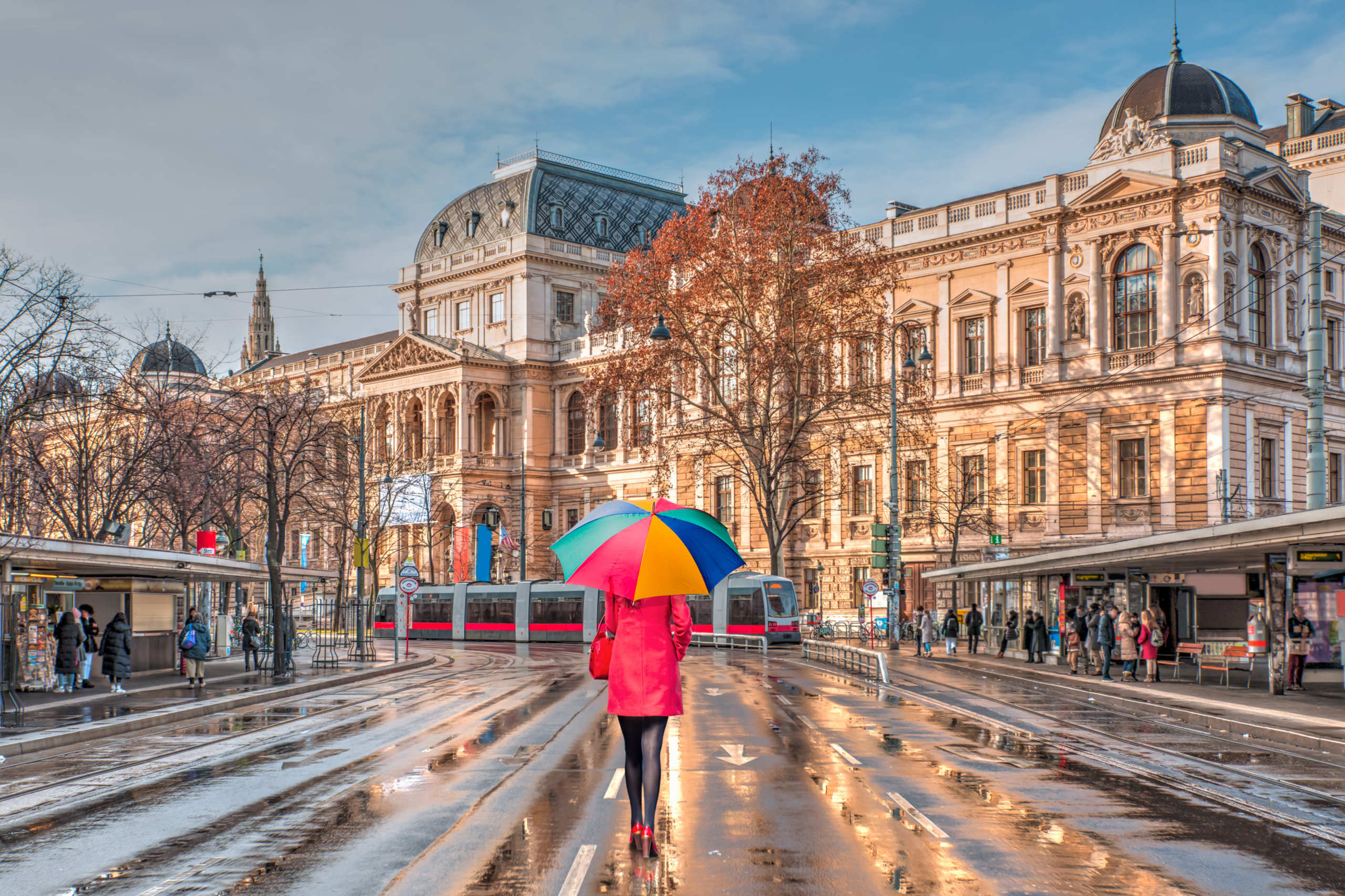 Vor der Universität in Wien steht eine Frau mit buntem Regenschirm im Regen. Symbolbild für die Kettenvertragsregelung.