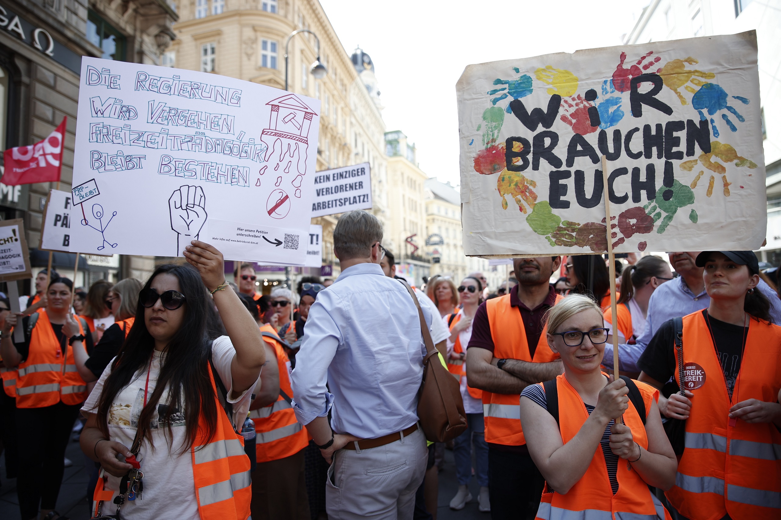 Demonstrierende in orangen Westen halten Schilder in die Luft. Auf einem Schild steht "Wir brauchen euch", auf dem anderen "Die Regierung wird vergehen, die Freizeitpädagogik wird bestehen."