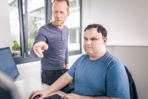 Martin Hartl zeigt mit seinem Zeigefinger auf einen Computer, Angelo sitzt an diesem Computer und hält die Maus. Symbolbild für Menschen mit Autismus im Arbeitsleben.