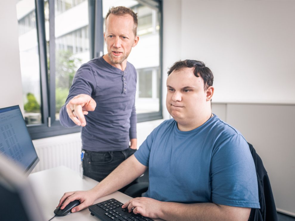 Martin Hartl zeigt mit seinem Zeigefinger auf einen Computer, Angelo sitzt an diesem Computer und hält die Maus. Symbolbild für Menschen mit Autismus im Arbeitsleben.