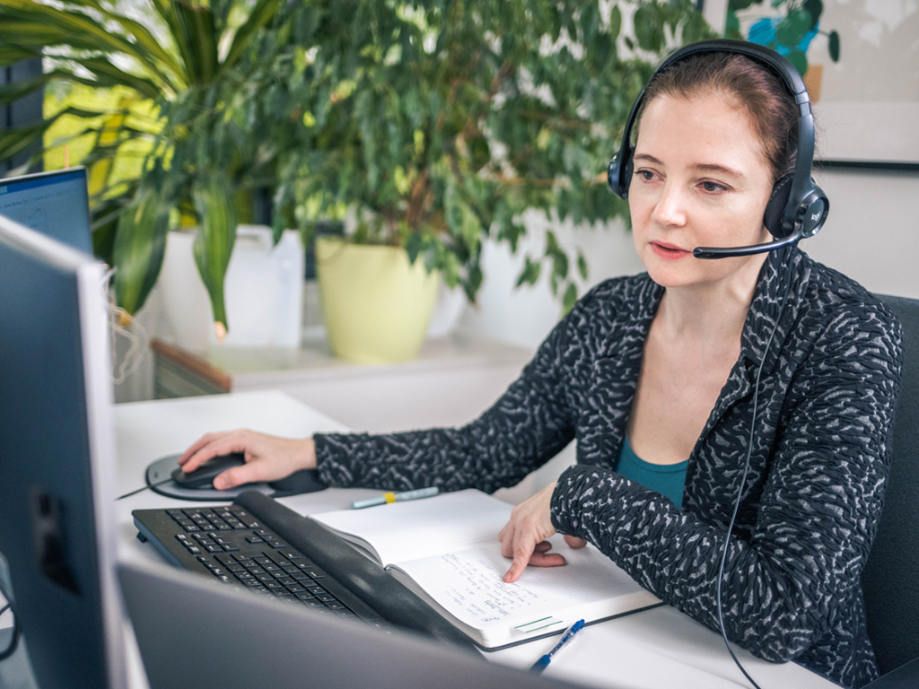 Beate Fabian sitzt vor ihrem Computer, hält in der linken Hand ihre Maus, unter sich ein Notizbuch. Sie trägt ein Headset. Fabian arbeitet mit Menschen mit Autismus zusammen.