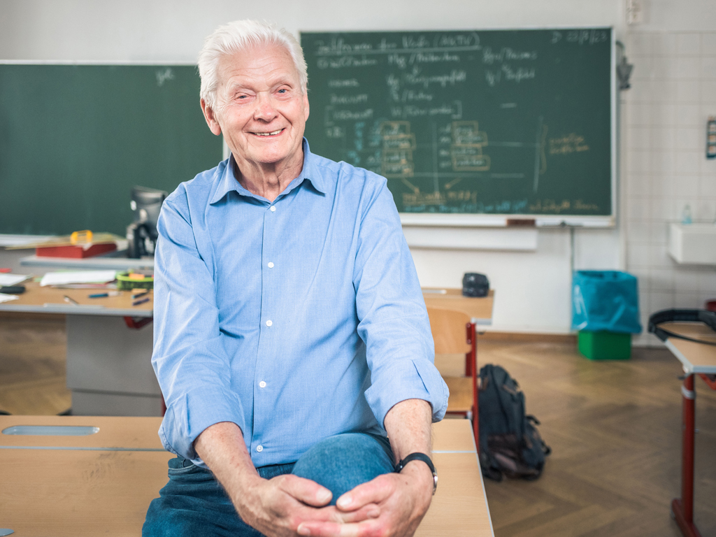 Reinhart Sellner sitzt auf einem Tisch vor einer beschriebenen Schultafel. Symbolbild für Pensionist:innen, die wieder in die Lohnarbeit zurückkehren.