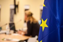 Eine Frau arbeitet an einem Schreibtisch. Im Vordergrund ist eine EU-Flagge zu sehen.