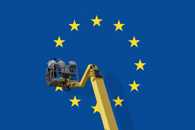 Eine EU-Flagge, an der auf einer Hebebühne zwei Arbeiter:innen arbeiten. Symbolbild für das soziale Europa.