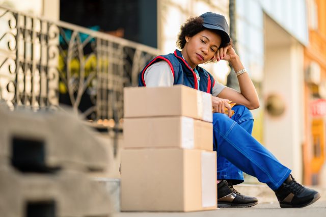 Eine Paketzustellerin sitzt vor Paketen. Sie wirkt müde. Symbolbild für Paketdienste und ihre Arbeitsbedingungen.