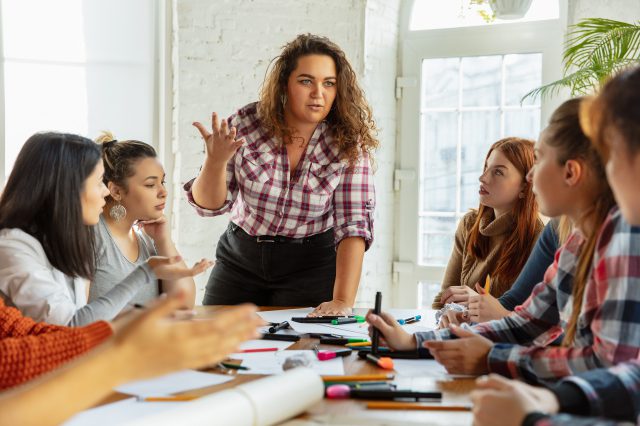 Junge Frauen sitzen an einem Tisch. Eine Frau steht und erklärt den anderen Frauen etwas. Symbolbild für Betriebsräte und ihre Herausforderungen, wenn Arbeitgeber ihren Betriebsrat verhindern wollen.