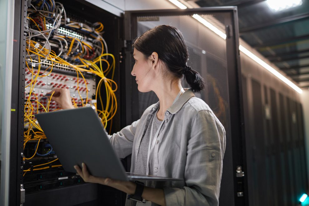 Eine Frau steht vor einem Server und verbindet Kabel. Sie hält einen Laptop in der Hand. Symbolbild für Digitalisierung am Arbeitsplatz.