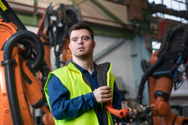 Ein junger Mann mit einer gelben Warnweste arbeitet an einem technischen Gerät. Er hat das Downsyndrom. Es ist ein Symbolbild für Jugendliche mit Behinderung, die jetzt Zugang zum AMS haben.