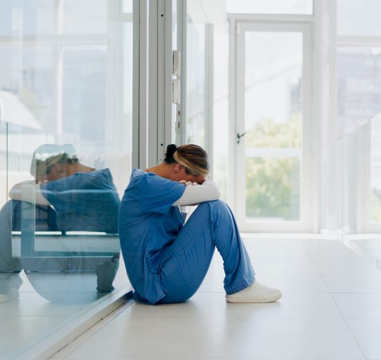 Eine Pflegerin sitzt auf dem Boden einer Klinik. Sie hat ihre Arme verschränkt, auf ihre Knie gelegt und ihr Blick geht nach unten. Symbolbild für die Pflegelehre.