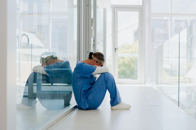 Eine Pflegerin sitzt auf dem Boden einer Klinik. Sie hat ihre Arme verschränkt, auf ihre Knie gelegt und ihr Blick geht nach unten. Symbolbild für die Pflegelehre.