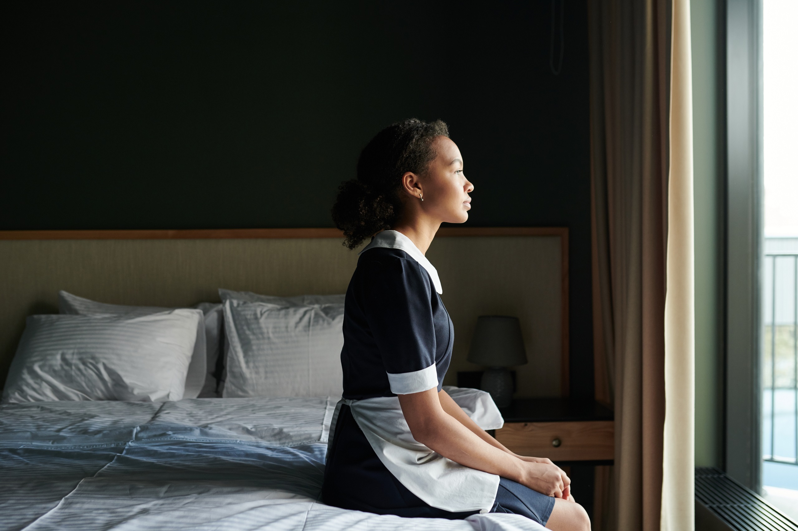 Eine Raumpflegerin sitzt auf einem Hotelbett und schaut müde nach draußen. Symbolbild für die Hotelbranche, die oft Betriebsräte verhindern lässt. 