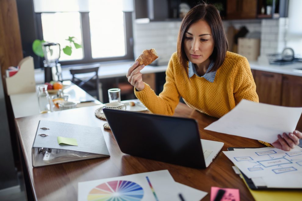 Eine Frau sitzt in ihrer Küche und arbeitet etwas an ihrem Laptop, während sie isst. vor- und Nachteile des Homeoffice.