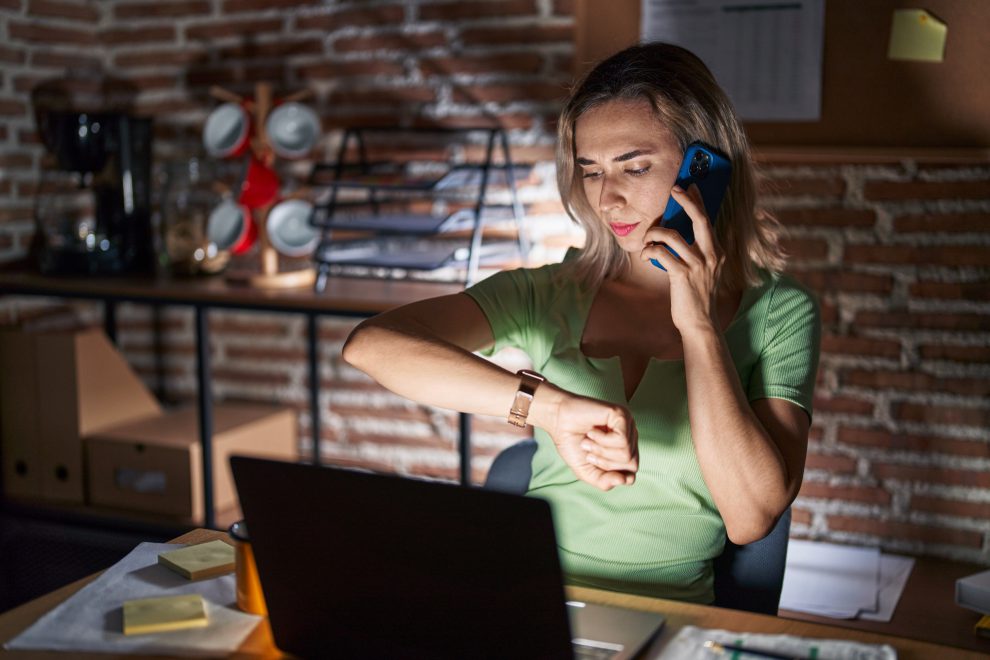 Eine Frau, die vor ihrem Laptop sitzt, schaut ihre Armbanduhr. Symbolbild für eine Arbeitszeitverkürzung.