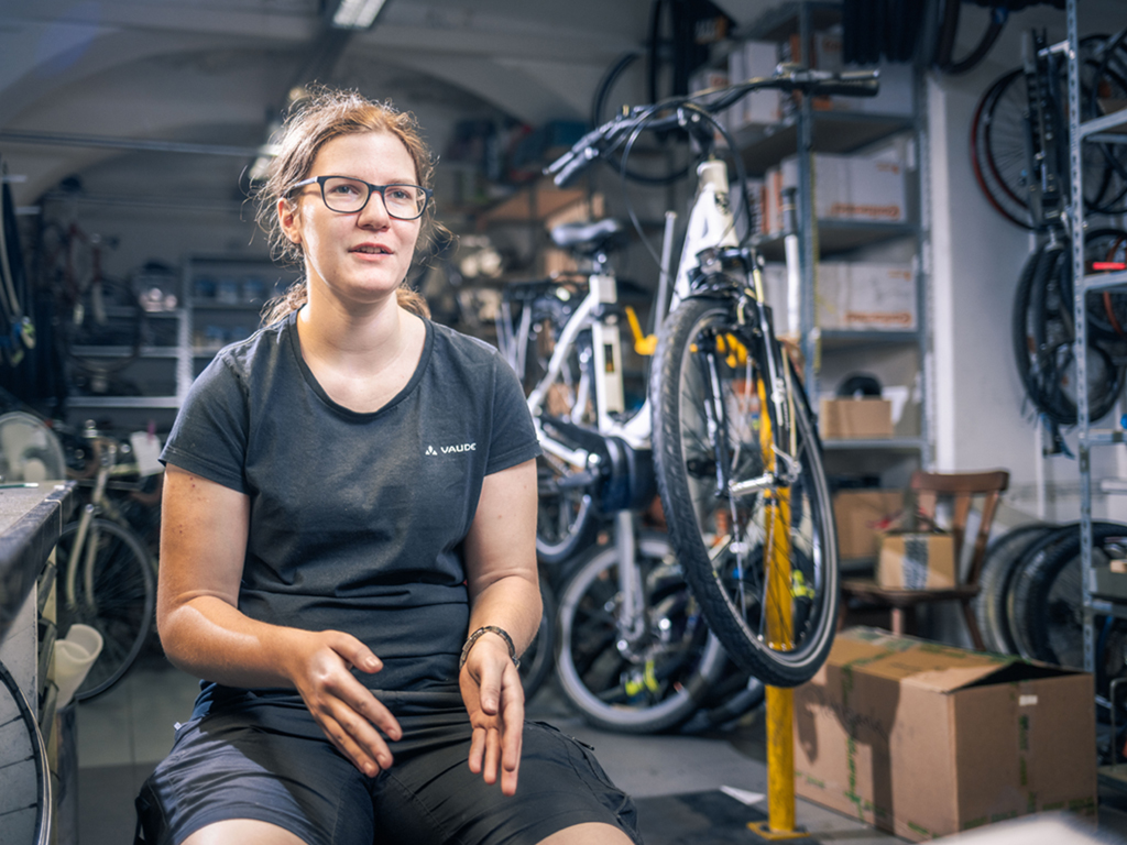 Sarah Sutlovic sitzt in der Fahrradwerkstatt, hinter ihr sind Fahrräder zu sehen. Sie spricht. Symbolbild für den Lehrberuf Fahrradmechatroniker:in.