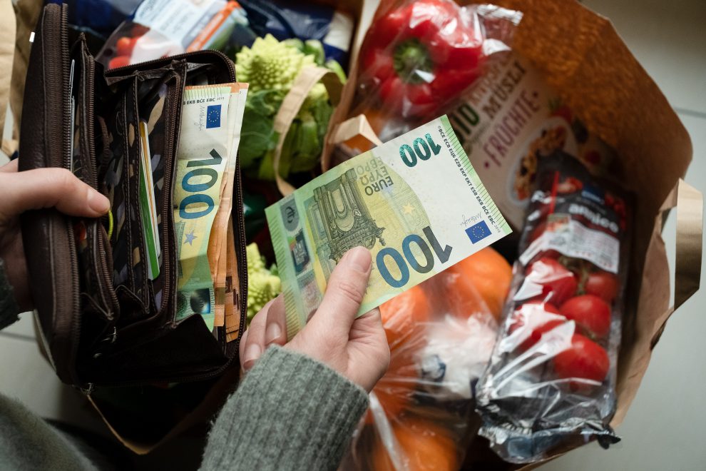 Eine volle Einkaufstüte. Davor hält eine Person eine Geldtasche in der Hand sowie einen 100-Euro-Schein. Symbolbild für die Teuerung.