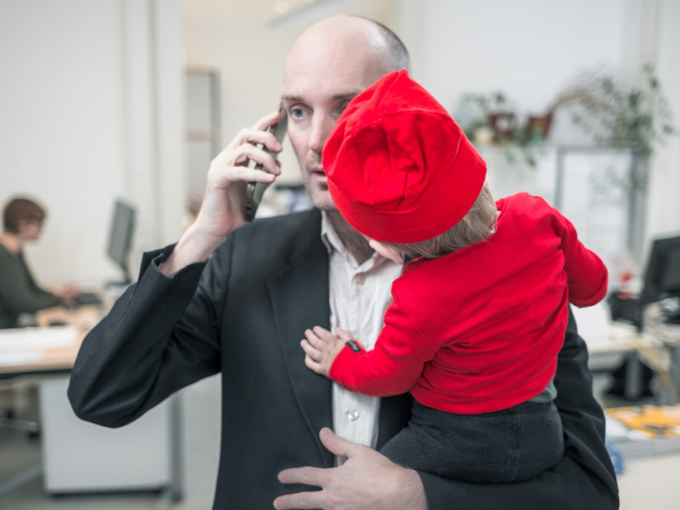 Ein Mann im Anzug telefoniert mit dem Handy arbeitet und hat dabei sein Kind auf dem Arm. Väter in Elternkarenz sind eine Minderheit.