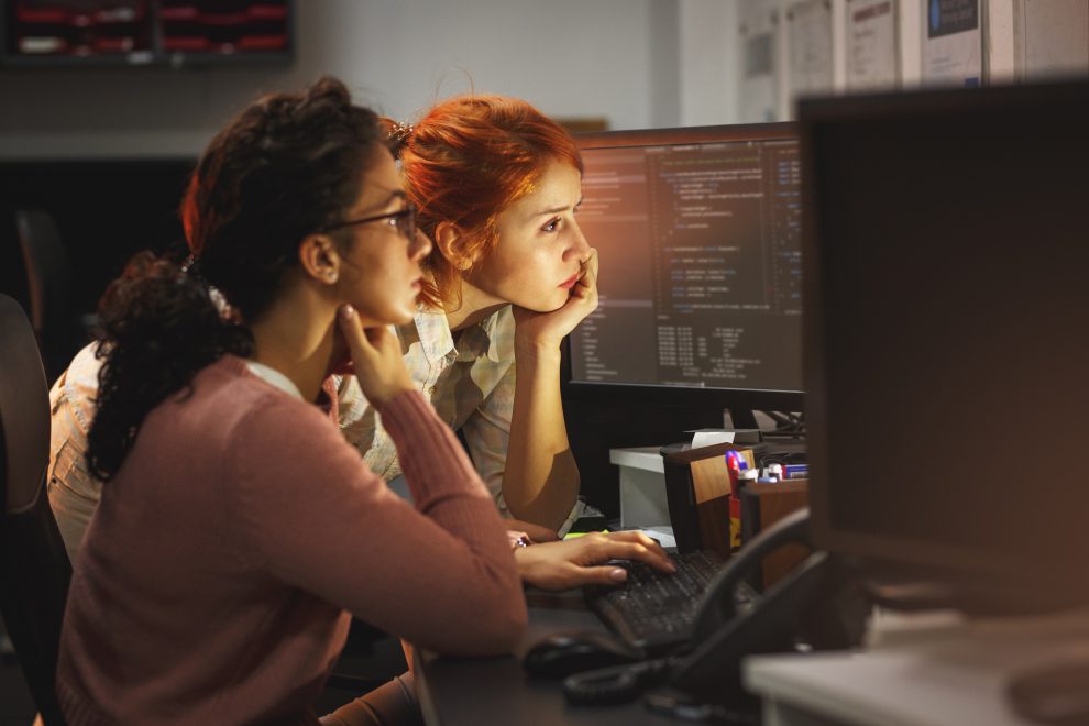 Zwei Frauen sitzen vor zwei Bildschirmen und programmieren. Symbolbild für Digitalisierung.