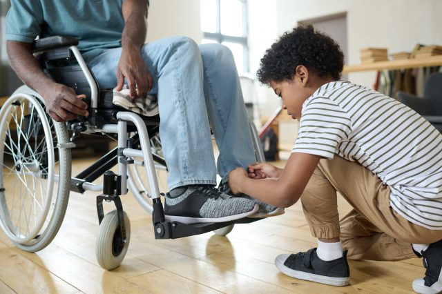 Ein Junge knotet seinem Vater die Schnürsenkel zu. Sein Vater sitzt im Rollstuhl. Symbolbild für junge Pflegende.