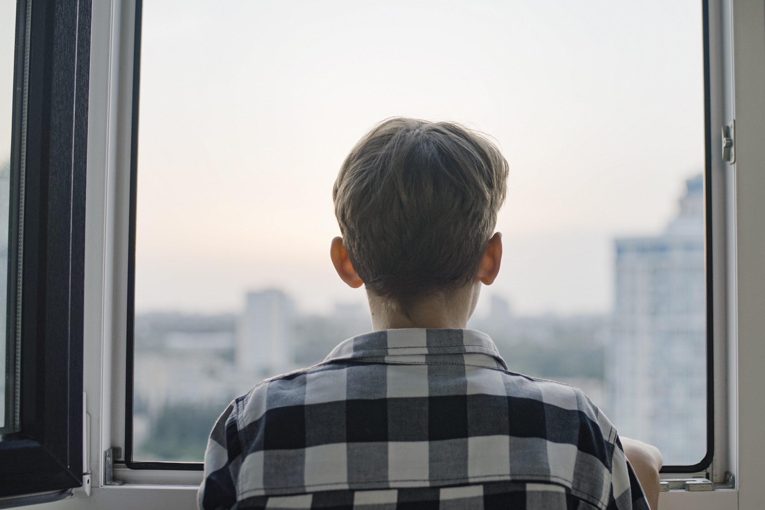 Kind schaut aus einem Fenster auf eine graue Stadt. Symbolbild für Kinderarmut.