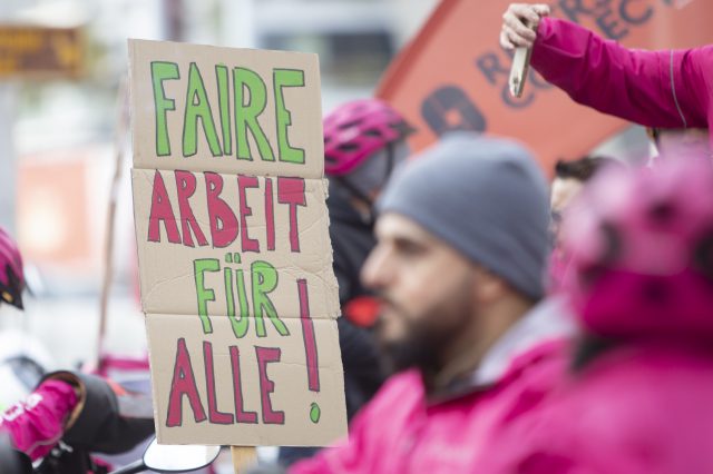 Ein Streik von Foodora-Ridern. Im Vordergrund ist ein Mann mit einer Trillerpfeife zu sehen. Im Hintergrund ein Schild mit der Aufschrift "Faire Arbeit für alle".