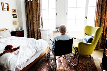 Eine Frau sitzt in einem Rollstuhl. Sie sitzt in einem Schlafzimmer, neben ihr ist ein Kuscheltier zu sehen sowie ein gelber Ohrensessel. Symbolbild für die Verschlechterungen in der Selbstverwaltung.